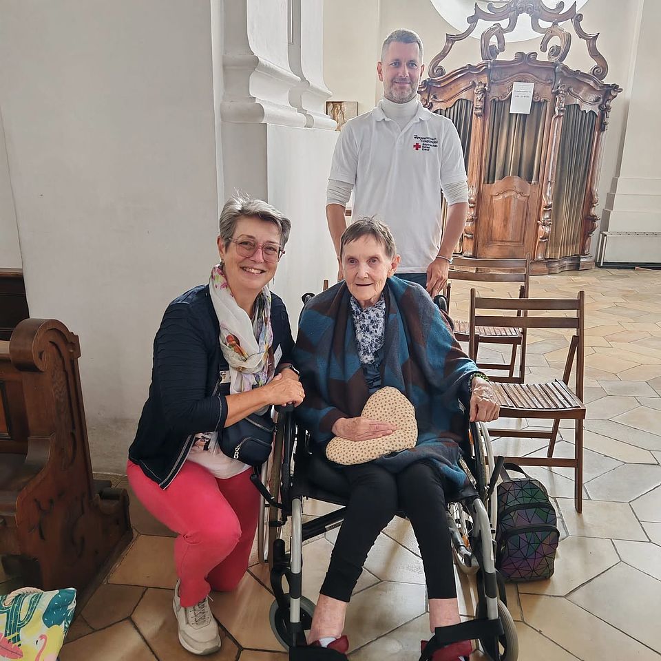 herzenswunschmobil_18102026_2 Das Foto zeigt zwei Damen in einer Kirche, hinter ihnen steht ein Beschäftigter des BRK Ingolstadt. Die ältere Dame sitzt im Rollstuhl.