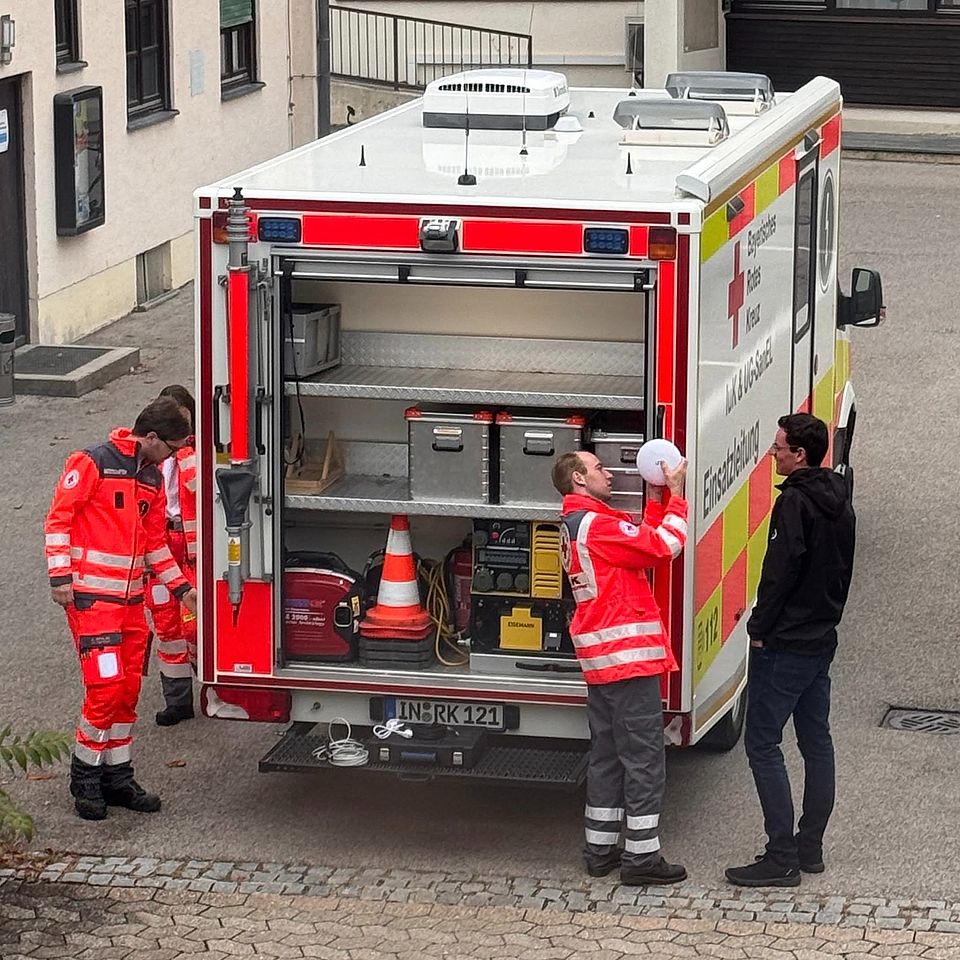 Das Foto zeigt die geöffenete Rückseite des Einsatzleitwagens. Davor stehen drei unifomierte Einsatzkräfte des BRK sowie einen zivil gekleiderten Mann auf dem BRK Gelände in Ingolstadt.