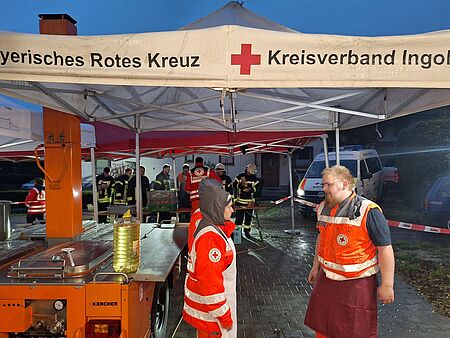 Das Foto zeigt eine orange-farbige Feldküche der Marke Kärcher unter einem Pavillon mit BRK-Aufdruck. Daneben stehen Köche in BRK-Uniform und hinter der Feldküche halten sich uniformierte Einsatzkräfte auf.
