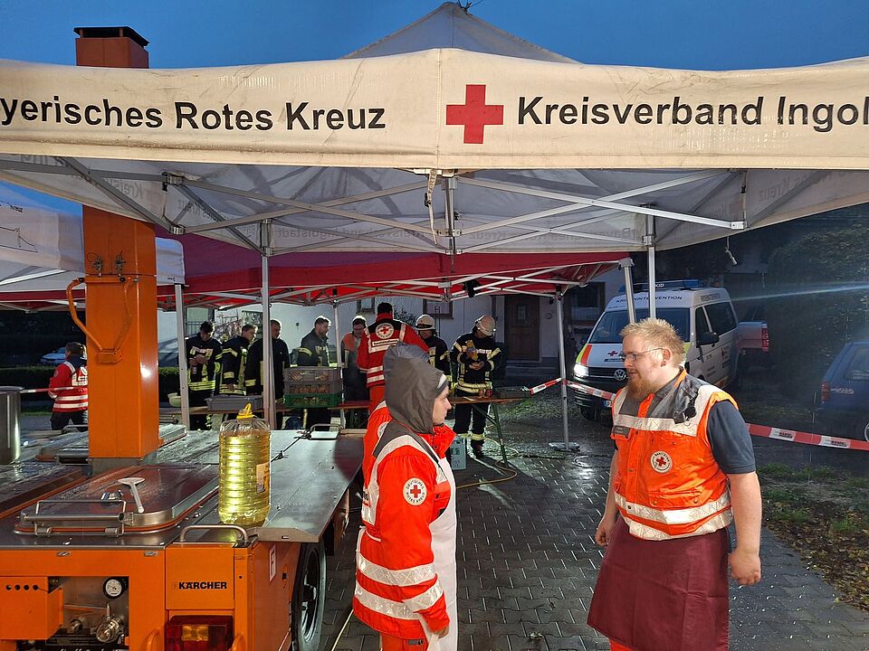 Das Foto zeigt eine orange-farbige Feldküche der Marke Kärcher unter einem Pavillon mit BRK-Aufdruck. Daneben stehen Köche in BRK-Uniform und hinter der Feldküche halten sich uniformierte Einsatzkräfte auf.