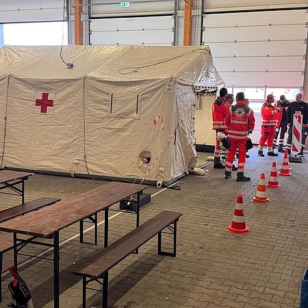 Das Foto zeigt ein weißes Zelt mit rotem Kreuz, rechts daneben stehen 7 Einsatzkräfte in roter Rot-Kreuz-Uniform.