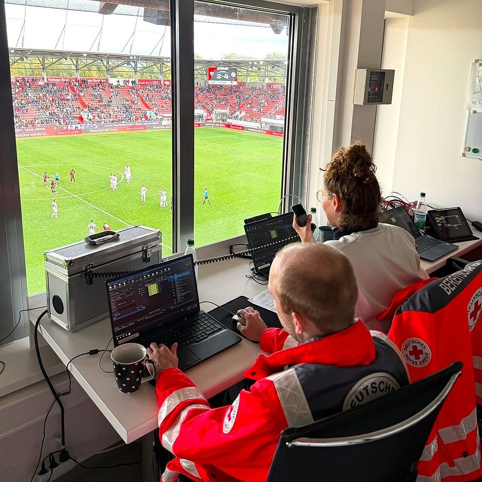 Das Foto zeigt zwei sitzende Einsatzkräfte, die von einer Kabine aus durch ein großes Fenster das Spiel des FC Ingolstadt beobachten.
